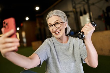 attraktive, reife Frau in Sportbekleidung mit Brille, die Selfies macht, während sie Hanteln hält