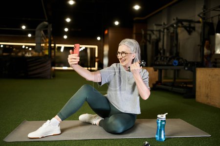 Photo for Positive mature sportswoman in gray t shirt with glasses taking selfies while holding dumbbells - Royalty Free Image