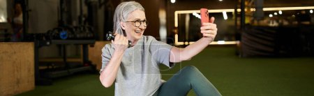 Téléchargez les photos : Femme sportive mature joyeuse en t-shirt gris avec des lunettes prendre des selfies tout en tenant des haltères, bannière - en image libre de droit