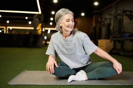 ansprechende reife, fröhliche Frau in bequemer Sportbekleidung sitzt auf dem Boden im Fitnessstudio und schaut weg