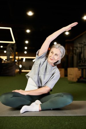 positive beautiful mature sportswoman in comfy attire sitting and stretching her muscles in gym