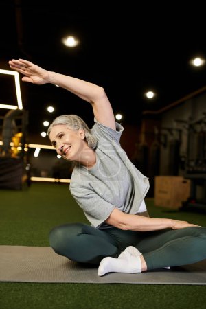 alegre atractiva mujer mayor en ropa deportiva cómoda con el pelo gris estirando sus músculos en el gimnasio