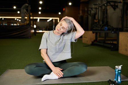Photo for Good looking athletic mature sportswoman in cozy attire sitting and stretching her muscles in gym - Royalty Free Image
