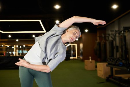 good looking jolly mature sportswoman in cozy outfit stretching her muscles while exercising in gym