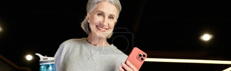 Téléchargez les photos : Femme mûre joyeuse avec cheveux gris tenant bouteille d'eau et téléphone et souriant à la caméra, bannière - en image libre de droit
