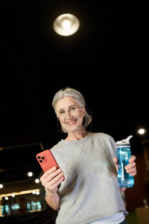 Photo for Cheerful mature sportswoman with gray hair holding water bottle and phone and smiling at camera - Royalty Free Image