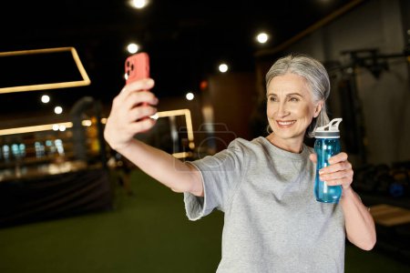 fröhliche gut aussehende Seniorin in kuscheliger Kleidung, die Selfies im Fitnessstudio macht und eine Wasserflasche in der Hand hält
