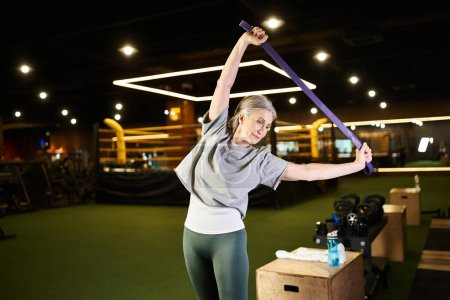 Photo for Positive mature pretty sportswoman in cozy attire training with fitness expander while in gym - Royalty Free Image