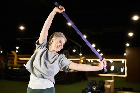 fröhliche reife Sportlerin in bequemer Kleidung, die im Fitnessstudio aktiv mit Fitness-Expander trainiert