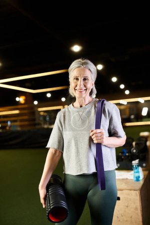 Photo for Cheerful mature sportswoman posing with fitness expander and weight bag and smiling at camera - Royalty Free Image