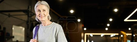 Photo for Positive senior woman posing in gym with fitness expander and weight bag and looking away, banner - Royalty Free Image