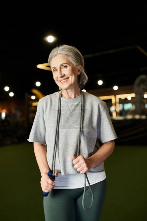 Foto de Deportista madura positiva con el pelo gris posando con saltar la cuerda y sonriendo a la cámara en el gimnasio - Imagen libre de derechos