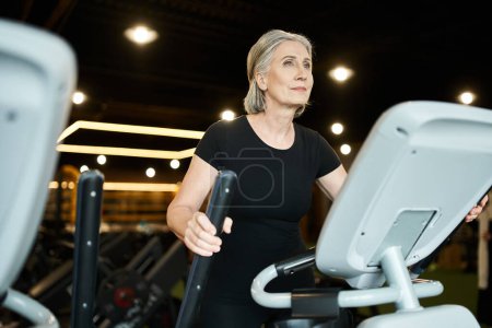 good looking jolly mature sportswoman in black t shirt exercising actively on elliptical trainer