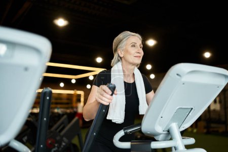 Foto de Mujer mayor deportiva positiva haciendo ejercicio en cross trainer mientras está en el gimnasio con toalla en los hombros - Imagen libre de derechos