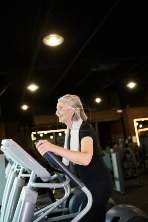 cheerful sporty mature woman exercising on cross trainer while in gym with towel on shoulders