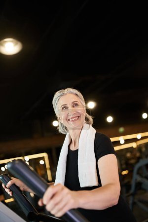 Foto de Mujer alegre bastante madura con pelo gris y toalla en los hombros haciendo ejercicio en cross trainer - Imagen libre de derechos