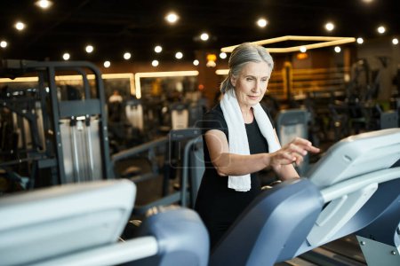 Photo for Beautiful mature sportswoman in black t shirt with towel on shoulders training on treadmill in gym - Royalty Free Image