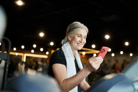 Téléchargez les photos : Joyeuse sportive mature aux cheveux gris ayant un appel vidéo pendant l'entraînement sur tapis roulant dans la salle de gym - en image libre de droit