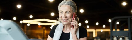 positive senior woman with towel on shoulders talking by phone while on treadmill in gym, banner