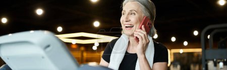 Foto de Mujer madura alegre con toalla en los hombros hablando por teléfono mientras en la cinta de correr en el gimnasio, pancarta - Imagen libre de derechos
