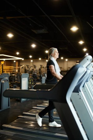 Foto de Mujer deportiva madura de buen aspecto en el entrenamiento de traje negro en la cinta de correr activamente mientras está en el gimnasio - Imagen libre de derechos