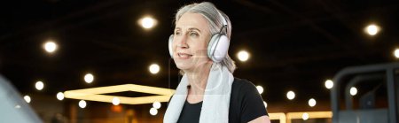 mature positive woman with gray hair and headphones exercising on treadmill in gym, banner