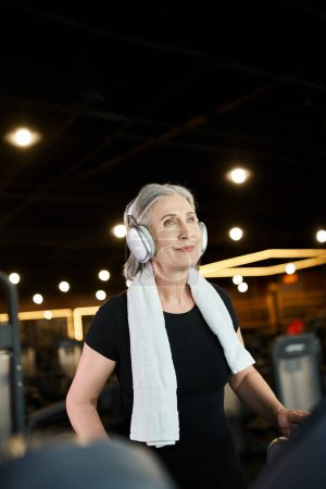 Foto de Atractiva mujer madura alegre con pelo gris y auriculares haciendo ejercicio en la cinta de correr en el gimnasio - Imagen libre de derechos