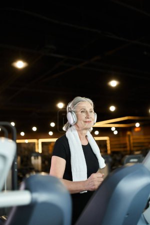 Foto de Alegre mujer mayor de pelo gris en entrenamiento de ropa deportiva en la cinta de correr y escuchar música en los auriculares - Imagen libre de derechos