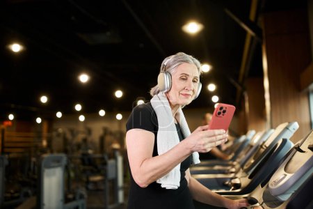 Foto de Mujer madura alegre con toalla en los hombros y auriculares sosteniendo el teléfono y haciendo ejercicio en la cinta de correr - Imagen libre de derechos