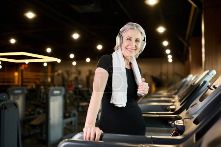 cheerful senior woman with towel and headphones smiling at camera while exercising on treadmill