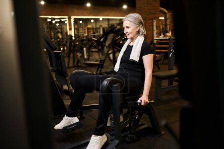 Foto de Atractiva deportista madura alegre con pelo gris y entrenamiento de toallas en la máquina de la pantorrilla mientras está en el gimnasio - Imagen libre de derechos