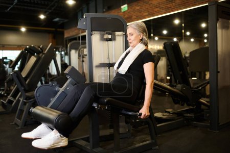 Foto de Atlética madura mujer de pelo gris en cómodo entrenamiento de ropa deportiva en la máquina de rizos de piernas mientras está en el gimnasio - Imagen libre de derechos