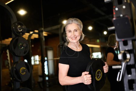 mujer madura alegre de buen aspecto con pelo gris sosteniendo el disco de peso y sonriendo a la cámara en el gimnasio