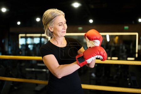 Téléchargez les photos : Séduisante femme mûre en vêtements de sport avec des cheveux gris et des gants de boxe posant sur l'anneau dans la salle de gym - en image libre de droit