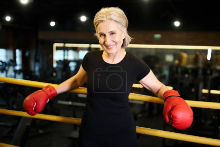 attractive joyous mature sportswoman with boxing gloves smiling at camera while on ring in gym