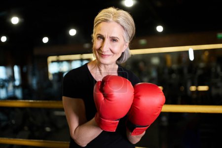 Foto de Atractiva deportista jubilosa con guantes de boxeo sonriendo a la cámara mientras está en el ring en el gimnasio - Imagen libre de derechos