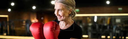 Foto de Mujer mayor bastante alegre en ropa deportiva con pelo gris y guantes de boxeo posando en el anillo, pancarta - Imagen libre de derechos