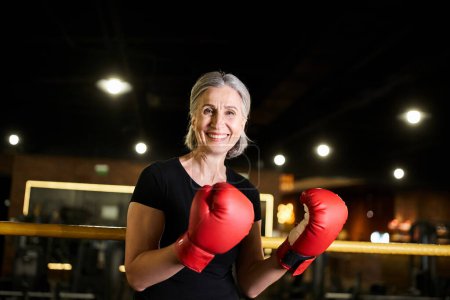 Foto de Atractiva mujer madura alegre en ropa deportiva con guantes de boxeo entrenamiento en el anillo y sonriendo a la cámara - Imagen libre de derechos
