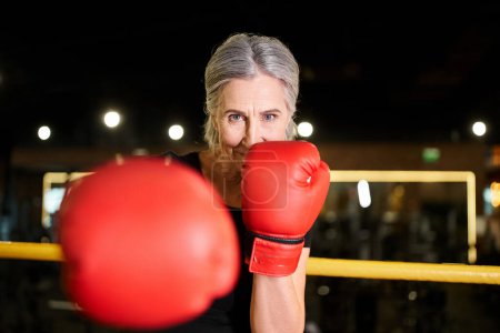 Téléchargez les photos : Belle femme sénior joyeux en vêtements de sport avec des gants de boxe de formation sur anneau et souriant à la caméra - en image libre de droit