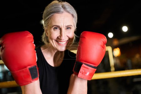 Téléchargez les photos : Séduisante femme âgée joyeuse en vêtements de sport avec des gants de boxe de formation sur anneau et souriant à la caméra - en image libre de droit