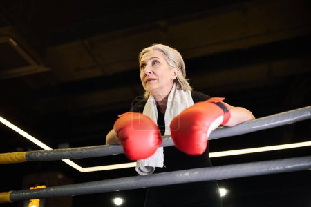 sportliche grauhaarige reife Frau mit Handtuch und Boxhandschuhen, die wegschaut, während sie im Fitnessstudio am Ring steht