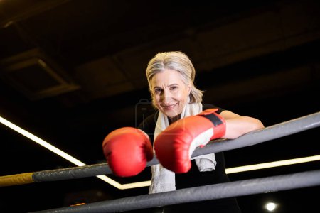 freudige athletische reife Frau mit Handtuch und Boxhandschuhen, die am Ring trainiert und in die Kamera lächelt