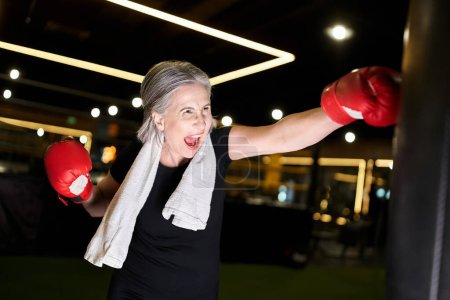 attraktive sportliche reife Frau mit grauen Haaren und Handtuch in Boxhandschuhen schlägt Boxsack in Turnhalle