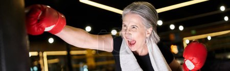 Photo for Pretty athletic mature sportswoman with towel and boxing gloves beating punching bag, banner - Royalty Free Image