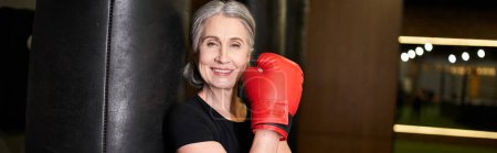 mature merry sportswoman with boxing gloves next to punching bag and smiling at camera, banner