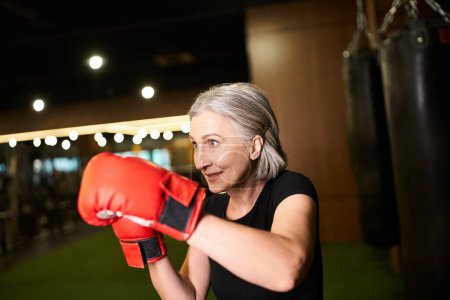 Foto de Atlética madura atractiva mujer con pelo gris posando con guantes de boxeo mientras entrena en el gimnasio - Imagen libre de derechos