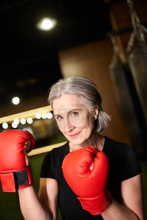Photo for Attractive senior merry woman in sportswear with gray hair posing with boxing gloves while in gym - Royalty Free Image