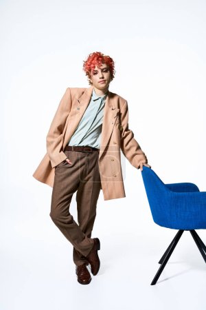 Mujer extraordinaria con el pelo rojo se para con confianza al lado de una silla.