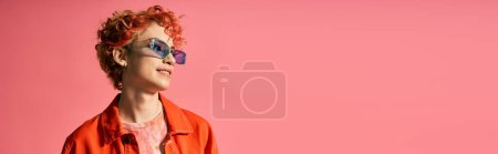 Foto de Una mujer elegante con el pelo rojo usando gafas de sol sobre un fondo rosa brillante. - Imagen libre de derechos