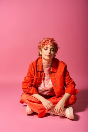 Foto de Una mujer llamativa con el pelo rojo está sentada sobre un vibrante fondo rosa. - Imagen libre de derechos